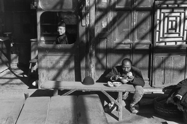 Henri Cartier-Bresson - Un camarero se sienta en la ventana abierta de una taberna, mientras que un "coolie" come afuera bajo una pérgola.